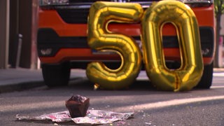 Zu sehen ist eine 50 aus Luftballons und ein Kuchen vor einem Auto.