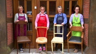 Vier Frauen in bunten Schürzen mit je einem alten Stuhl vor sich.