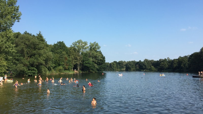 Blick von der Brücke, die über den Achterdieksee führt in Richtung des weiten Teils des Sees. Badende stehen versprenkelt im Wasser.