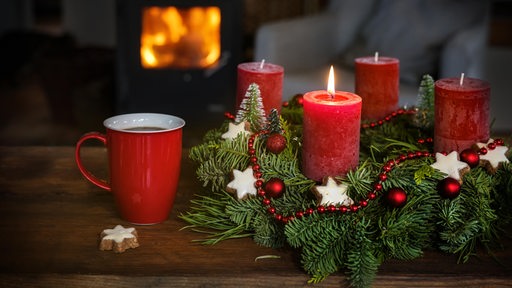 Ein Adventskranz mit Weihnachtsdekoration und roten Kerzen sowie eine Tasse Kaffee stehen auf einem Tisch vor dem Kamin in einem Wohnzimmer.