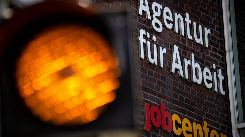 Eine Ampel leuchtet vor dem Schild der Agentur für Arbeit und des Jobcenter.