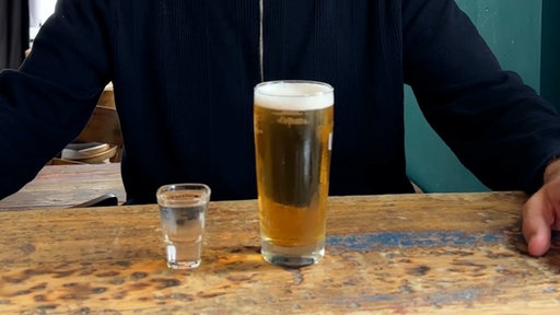 Ein Mann sitzt an einem Tresen vor einem Glas Bier und einem kleinen Schnapsglas mit farblosem Inhalt.