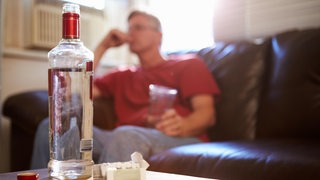 Auf einem Wohnzimmertisch steht eine Flasche Schnaps, im Hintergrund ein Mann mit Glas auf dem Sofa.