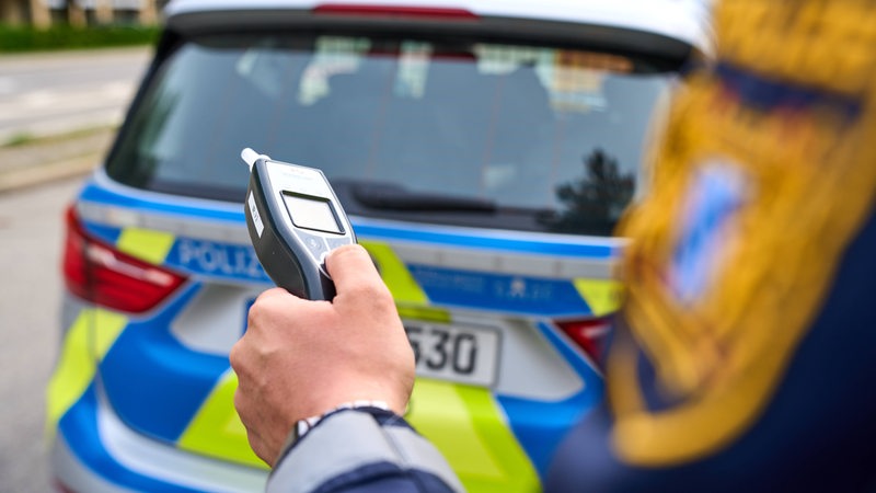 Ein Polizist steht hinter einem Streifenwagen und hält ein Alkoholmessgerät in der Hand.