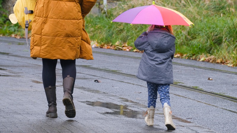 Eine Frau, die einen Kinderrucksack trägt, und ein Kind, das einen aufgespannten Regenschirm über dem Kopf hält, gehen eine mit Pfützen versehene Straße entlang.