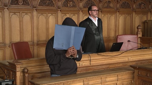 In einem Gerichtssal steht ein Richter und der Angklagte, welcher sich sein Geschicht hinter einer Mappe versteckt.
