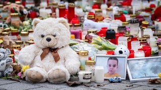 Ein Teddybär und ein Foto eines kleinen Jungen stehen im Vordergrund. Dahinter sind zahlreiche Kerzen zu sehen.