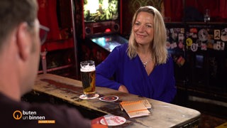 Eine Frau mit mittellangen blonden Haaren und einem blauen Kleid sitzt in einer Kneipe auf einem Barhocker mit einem Bier auf dem Tisch. 