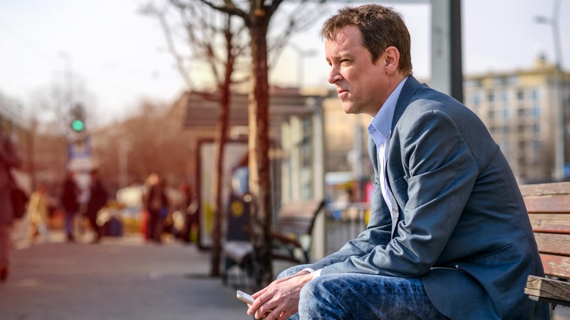 Ein Mann sitzt mit Handy in der Hand auf einer Bank und schaut nachdenklich.