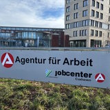 Ein Schild mit der Aufschrift "Agentur für Arbeit" steht auf einer Wiese vor mehreren Gebäuden.