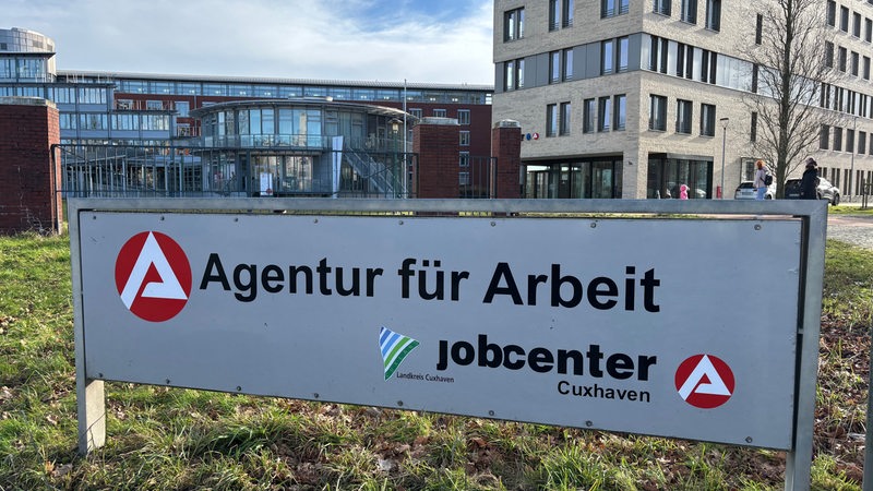 Ein Schild mit der Aufschrift "Agentur für Arbeit" steht auf einer Wiese vor mehreren Gebäuden.