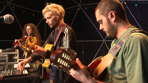 Drei Mitglieder einer Band stehen während eines Soundchecks auf einer Bühne.