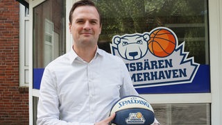 Der neue Geschäftsführer der Basketballer Eisbären Bremerhaven, Johannnes Marggraf, steht lächelnd mit einem Basketball in der Hand vor der Geschäftsstelle.