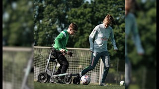 Ein Junge mit dunklen Haaren und Brille an einem Rollator spielt gemeinsam mit einem Mädchen Fußball.