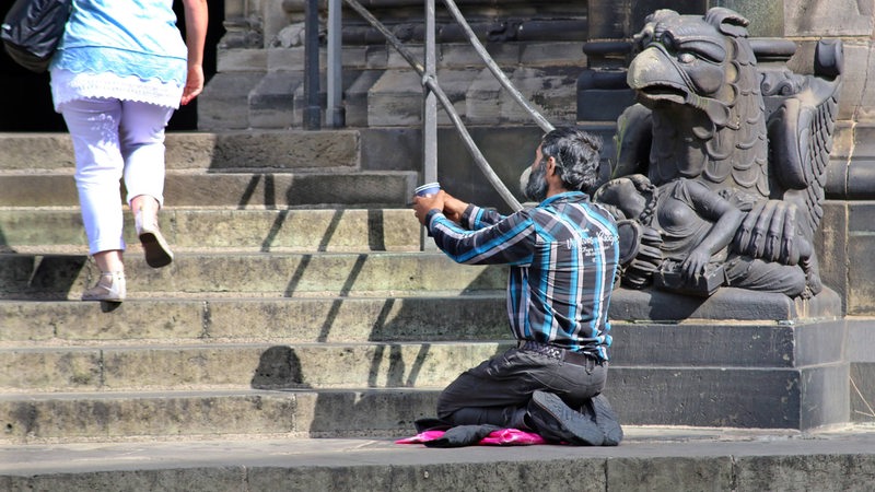 Ein Mann kniet vor den Stufen des Doms und hält einen Becher in die Höhe.