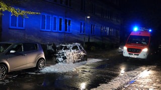 Vor einem Wohnhaus in Bremen-Gröpelingen stehen ein ausgebranntes Auto und ein Feuerwehr-Rettungswagen mit Blaulicht.