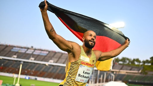 Der Bremer Weitspringer Leon Schäfer läuft lächelnd mit einer Deutschland-Fahne hinter dem Rücken durch ein Leichtathletik-Stadion.