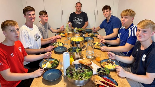 Mehrere junge Sportler sitzen um einen Esstisch vor gefüllten Tellern und lächeln.