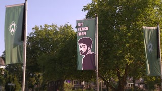 Eine Fahne weht vor dem Weser-Stadion mit einer Portät-Zeichnung der Hamas-Geisel Hersh Goldberg-Polin mit der Aufschrift "Let Hersh free!". 