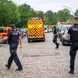 Drei Polizisten riegeln eine Straße ab, in der ein Wagen der Feuerwehr steht.