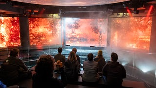 Museumsmitarbeiter und andere Personen sitzen auf der Erlebnisplattform während einer Besichtigung der Ausstellung zum Thema Wetterextreme im Klimahaus in Bremerhaven.