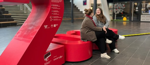 Zwei Personen sitzen auf einer roten Installation in einer Halle.