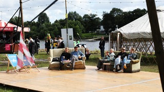 Auf einer Holzbühne neben der Weser sitzen Menschen auf Sofas.