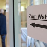 Menschen gehen in einer Grundschule an einem Hinweis mit der Aufschrift "Zum Wahlraum" vorbei,