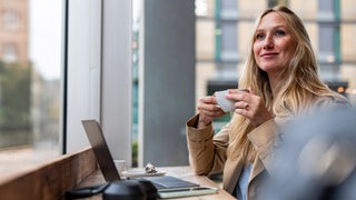 Eine blonde Frau sitzt in eine, Café vor ihrem Laptop, hält eine Kaffeetasse in der Hand und schaut in die Ferne.