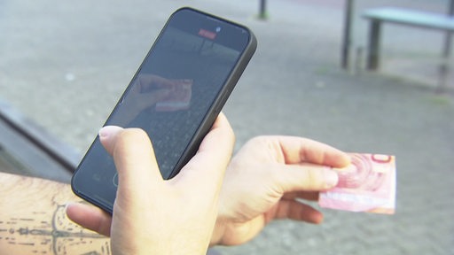 Ein Mann hält in einer Hand ein Handy, in der anderen einen 10-Euro-Schein.