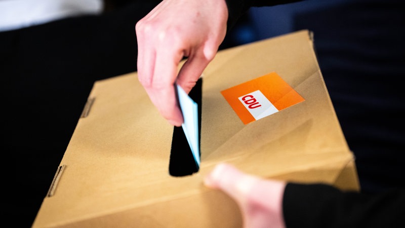 Eine Hand steckt einen Stimmzettel in einen Karton mit einem CDU-Aufkleber.