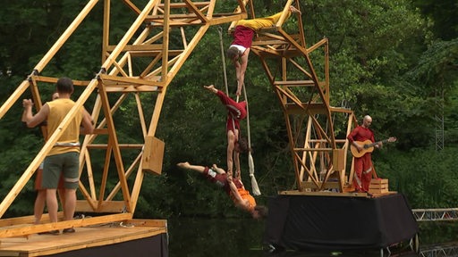 An einer Holzbrücke hängen drei Akrobaten und performen ein Kunststück. 