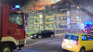 Ein brennendes Mehrfamilienhaus im Hintergrund, davor ein Feuerwehrwagen.