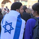 Ein Mann steht mit dem Rücken zur Kamera und trägt eine Israel-Flagge um seine Schultern. Eine Frau steht neben ihm und unterhält sich mit ihm.