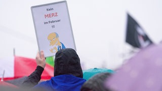 Ein Demonstrant hält ein Schild hoch, auf dem der Simpsons-Charakter Montgomery Burns zu sehen ist sowie der Text: "Kein Merz im Februar!!!"