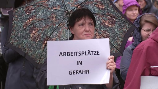 Eine Frau steht unter einem Schirm und hält ein Schild hoch mit der Aufschrift: "Arbeitsplätze in Gefahr"