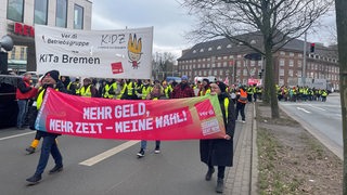 Menschen in Warnwesten tragen bei einer Demo verdi-Transparente und laufen über eine Straße.