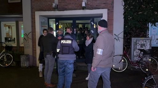 Mehrere Demonstrierende stehen vor einem Eingang und blockieren einen Polizisten vom Eintreten