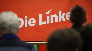 Menschen sitzen vor einem Logo mit der Aufschrift "Die Linke"