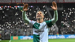 Diego reißt die Hände in die Luft. Im Hintergrund leuchten Handylichter auf den Rängen im Stadion.