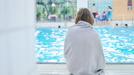 Ein Mädchen sitzt am Rand eines Schwimmbeckens und ist mit einem Handtuch bedeckt.