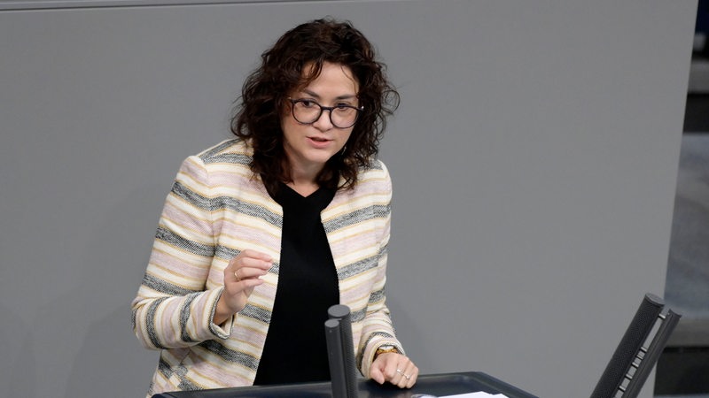 Eine Frau mit dunklen lockigen halblangen Haaren und Brille steht an einem Rednerpult mit der Aufschrift deutscher Bundestag und hebt den rechten Arm leicht. 