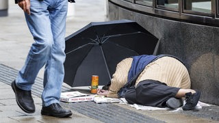 Eine obdachlose Person liegt schlafend auf dem Bürgersteig, eine Person läuft vorbei (Archivbild).