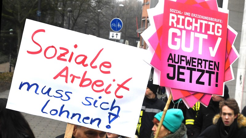 Mitglieder der Gewerkschaft für Erziehung und Wissenschaft (GEW) und Verdi halten in Bremen Plakate mit "Soziale Arbeit muss sich Lohnen!" und "Richtig Gut Aufwerten Jetzt" auf einer Kundgebung hoch. (Archivbild)