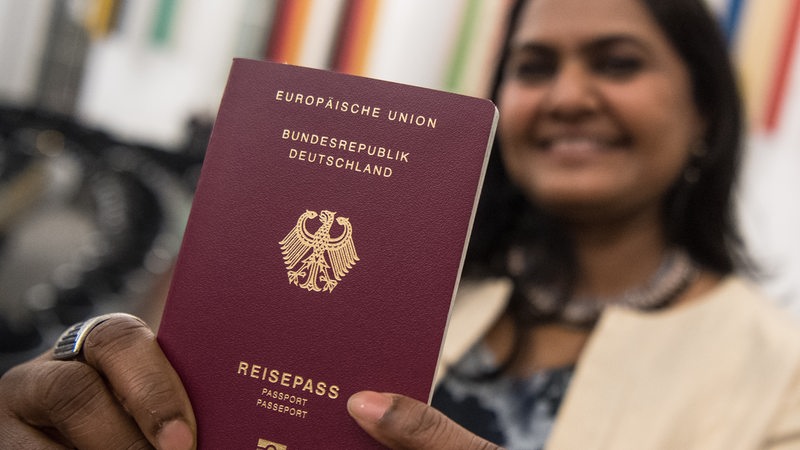 Eine aus Indien stammende junge Frau hält während einer Einbürgerungsfeier lachend ihren deutschen Reisepass in die Kamera.