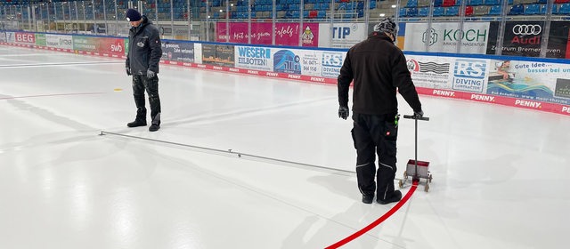 Ein Arbeiter zieht mit einem kleinen Farbwagen an der Hand den roten Kreis auf die Eisfläche in der Eisarena Bremerhaven.