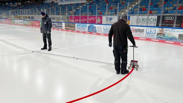 Ein Arbeiter zieht mit einem kleinen Farbwagen an der Hand den roten Kreis auf die Eisfläche in der Eisarena Bremerhaven.