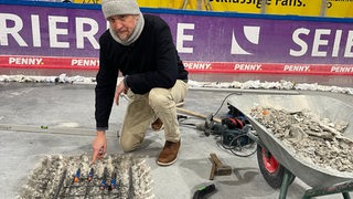 Stadthallenchef Gimpel zeigt auf ein Loch im Boden der Eisarena Bremerhaven. Neben ihm steht eine Schubkarre.