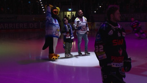 Ein Kind steht neben einem Maskottchen und einer weiteren Person im Scheinwerferlicht in einer Eisarena.