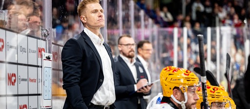 Alexander Sulzer, Cheftrainer des Eishockey-Teams der Fischtown Pinguins, steht mit ernster Miene an der Bande, vor ihm sitzen einige Ersatzspieler.